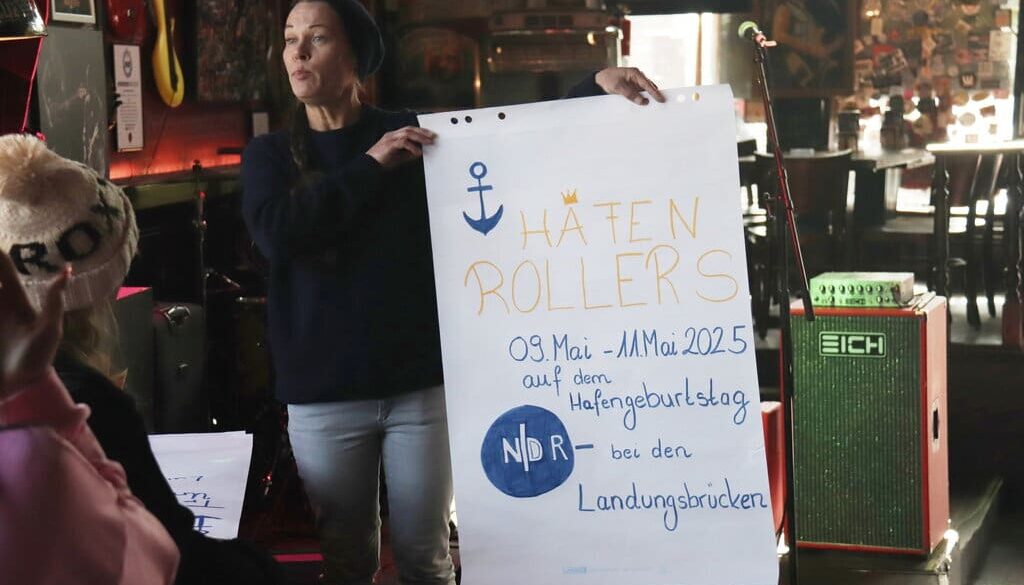 Gruppenleitung Vanessa hält ei A1 Plakat mit der Überschrift HAFENROLLERS und Infos zum inklusiven Hafengeburtstag hoch