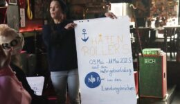 Gruppenleitung Vanessa hält ei A1 Plakat mit der Überschrift HAFENROLLERS und Infos zum inklusiven Hafengeburtstag hoch