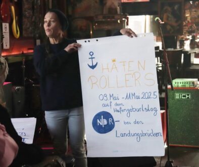 Gruppenleitung Vanessa hält ei A1 Plakat mit der Überschrift HAFENROLLERS und Infos zum inklusiven Hafengeburtstag hoch