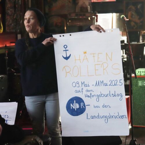 Gruppenleitung Vanessa hält ei A1 Plakat mit der Überschrift HAFENROLLERS und Infos zum inklusiven Hafengeburtstag hoch