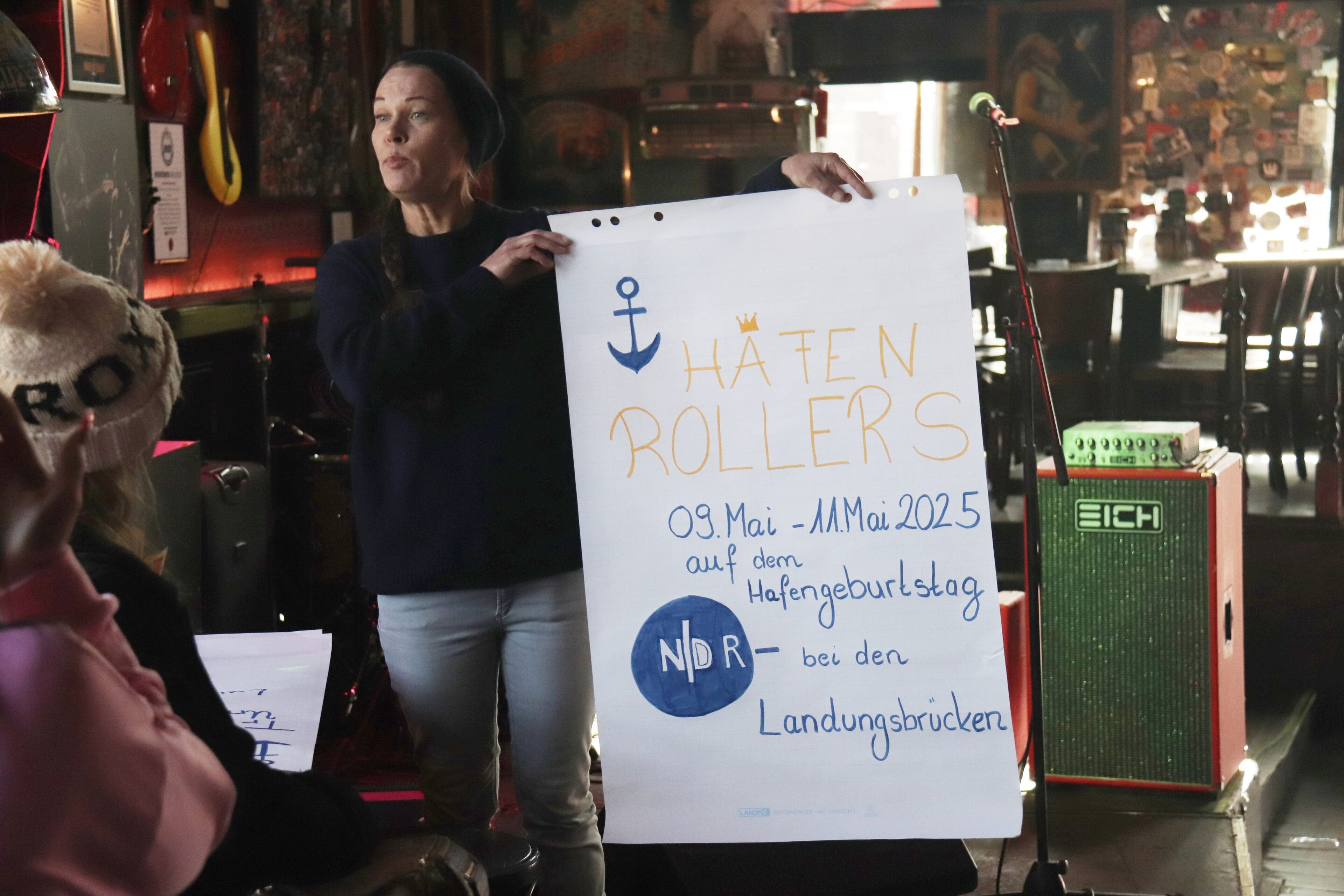 Gruppenleitung Vanessa hält ei A1 Plakat mit der Überschrift HAFENROLLERS und Infos zum inklusiven Hafengeburtstag hoch